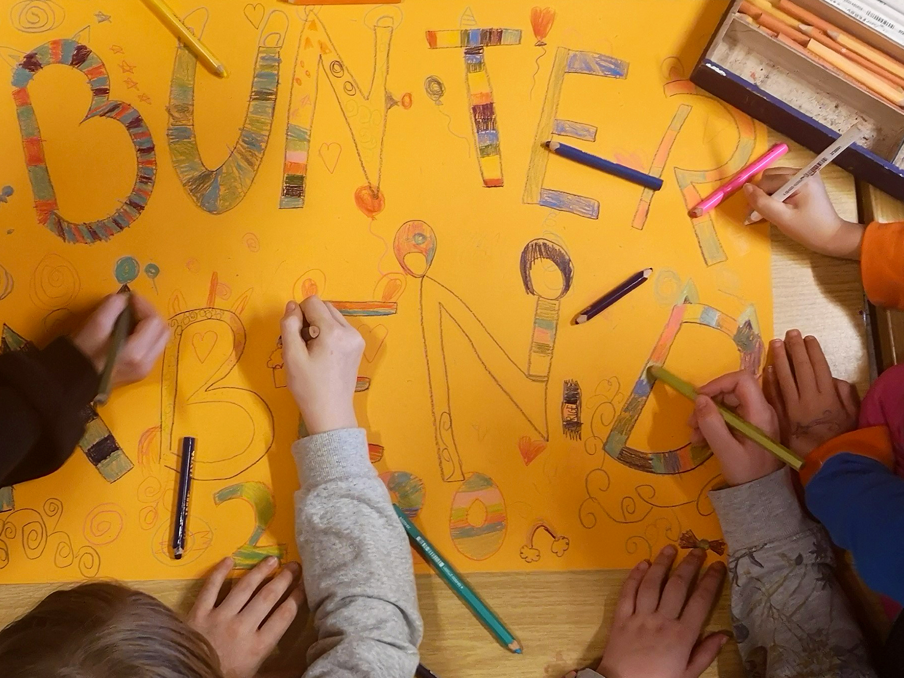 Kinder gestalten gemeinsam ein buntes Plakat. Man sieht viele Hände mit Buntstiften.